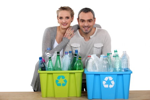Two individuals, a woman with blonde hair tied back and a man with dark short hair, are positioned behind a wooden surface, smiling at the camera. In front of them are two plastic recycling bins, one green and one blue, each featuring a white recycling symbol on the front. The green bin contains various empty plastic bottles in different sizes and shapes, with some caps visible, and the bottles are mostly transparent or light green. The blue bin also holds an assortment of plastic bottles, some with labels or caps, and both bins are situated outdoors on a paved or concrete area, with a plain white background, indicating a professional setting for rubbish or waste management services. The scene suggests a focus on waste sorting and recycling, in line with the services provided by Commercial Waste Sydenham.