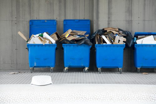The image depicts a row of four large blue wheelie bins positioned on a concrete pavement against a plain, smooth grey concrete wall. The first bin on the left is open, revealing a mix of white and green cardboard packaging, along with some plastic items, and is partially filled with paper and cardboard waste. The second bin is also open, filled with a variety of flattened cardboard boxes and paper, with some overhanging the edges. The third bin appears to contain mostly flattened cardboard and paper materials inside a black plastic bag, which is slightly pulled over the edge of the bin. The fourth bin is partially obscured, with small amounts of debris visible near the top. The pavement in front of the bins is clean, with a small white object, possibly a paper or cloth, lying on the ground near the left side of the image. The scene suggests an outdoor waste collection area, typical of commercial or residential rubbish disposal, consistent with rubbish removal services in the Sydenham area, as provided by Commercial Waste Sydenham.