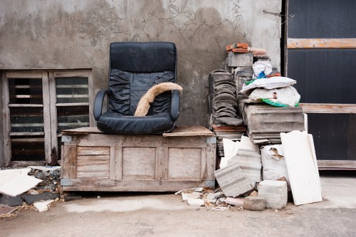 The image depicts a cluttered outdoor scene in a commercial or industrial area, with a black office chair featuring a plush, beige cushion on the seat positioned on top of a weathered wooden crate or storage box. To the right of the chair, there is a stack of grey concrete bricks and blocks, some with worn or chipped edges, piled haphazardly on the pavement. Several white and grey panels, possibly made of foam or plasterboard, are leaning against the brick pile, with additional debris and broken pieces scattered on the ground nearby. The background includes a beige wall with a small vent and a narrow grate, and the scene appears to be located in a back alley or side yard, indicative of waste removal or clearance activities typical of services by Commercial Waste Sydenham in the South London area. The environment is utilitarian, with visible weathering and signs of previous use, emphasizing the need for professional rubbish removal in this setting.
