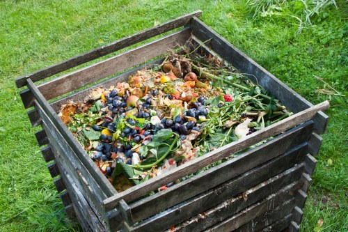 A rectangular wooden waste bin with dark weathered planks, positioned outdoors on a grassy surface, filled with a mixture of biodegradable garden waste and food scraps including vegetable peelings, fruit skins, and plant trimmings. The bin's slatted sides are visible, and the contents are overflowing slightly, showing a variety of textures and colours such as green leaves, brown peels, and bits of fruit. Surrounding the bin is a well-maintained green lawn, with no other objects or structures immediately nearby, reflecting a typical garden setting suitable for waste collection and removal services by companies like Commercial Waste Sydenham in the Sydenham area.