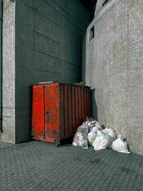 A large, rusted red metal dumpster with a weathered finish is positioned against the corner where two grey building walls meet, one with smooth stone cladding and the other with a textured concrete su