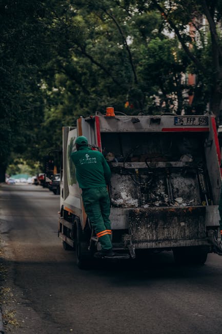 A sanitation worker dressed in a green uniform with reflective orange stripes is standing next to a small, grey and white rubbish collection vehicle on an urban street. The worker is positioned at the