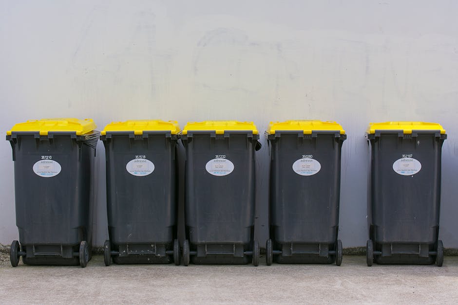 A sanitation worker dressed in a green uniform with reflective orange stripes is standing next to a small, grey and white rubbish collection vehicle on an urban street. The worker is positioned at the