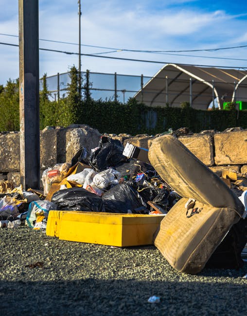 Three large black plastic bags filled with waste are positioned on a concrete pavement near a metal fence with vertical bars. The bags appear to contain household or garden rubbish, with some visible 